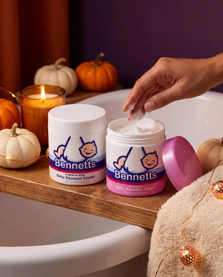 A hand scooping Bennetts Fragranced Aqueous Cream from the tub on a bath caddy, beside a Fragrance-Free tub, a candle and little pumpkins