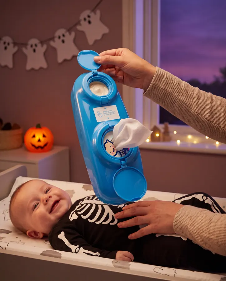 Hands opening a blue Bennetts Bum Buddy over a smiling baby in a skeleton onesie, with ghost bunting behind